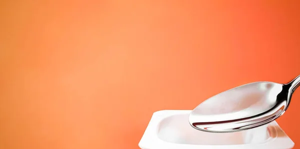 Tazza di yogurt e cucchiaio d'argento su fondo arancione, contenitore di plastica bianca con crema di yogurt, latticini freschi per una dieta sana e un equilibrio nutrizionale — Foto Stock