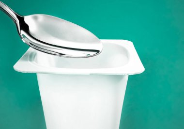 Yoğurt kabı ve yeşil arka planda gümüş kaşık yoğurt kremalı beyaz plastik kap sağlıklı beslenme ve beslenme dengesi için taze süt ürünü.