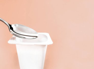 Yoğurt kabı ve bej arka planda gümüş kaşık, yoğurt kremalı beyaz plastik kap, sağlıklı beslenme ve beslenme dengesi için taze süt ürünü