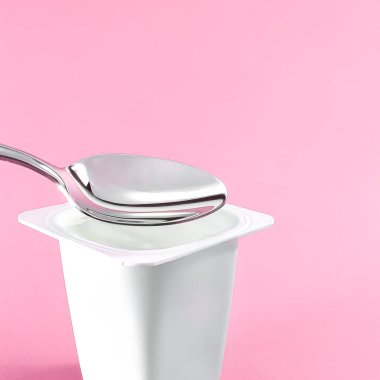 Yoğurt kabı ve pembe arka planda gümüş kaşık yoğurt kremalı beyaz plastik kap sağlıklı beslenme ve beslenme dengesi için taze süt ürünü.