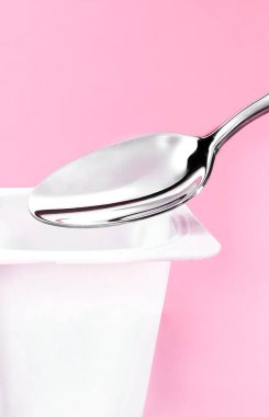 Yoğurt kabı ve pembe arka planda gümüş kaşık yoğurt kremalı beyaz plastik kap sağlıklı beslenme ve beslenme dengesi için taze süt ürünü.