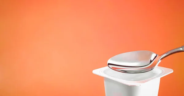 Tazza di yogurt e cucchiaio d'argento su fondo arancione, contenitore di plastica bianca con crema di yogurt, latticini freschi per una dieta sana e un equilibrio nutrizionale — Foto Stock