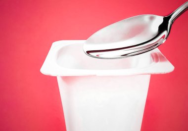 Kırmızı arka planda yoğurt kabı ve gümüş kaşık yoğurt kremalı beyaz plastik kap sağlıklı beslenme ve beslenme dengesi için taze süt ürünü.