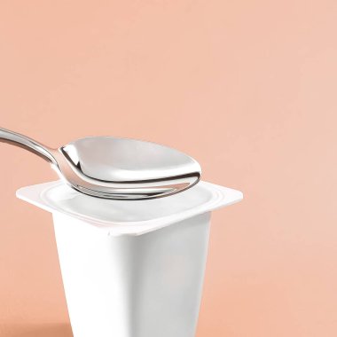 Yoğurt kabı ve bej arka planda gümüş kaşık, yoğurt kremalı beyaz plastik kap, sağlıklı beslenme ve beslenme dengesi için taze süt ürünü
