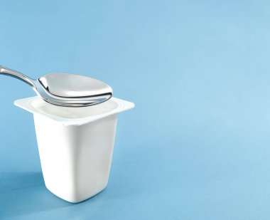 Yoğurt kabı ve mavi arka planda gümüş kaşık, beyaz plastik kap, sağlıklı beslenme ve beslenme için taze süt ürünü