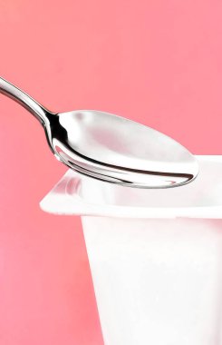 Yoğurt kabı ve pembe arka planda gümüş kaşık yoğurt kremalı beyaz plastik kap sağlıklı beslenme ve beslenme dengesi için taze süt ürünü.