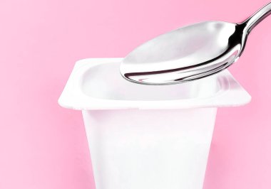 Yoğurt kabı ve pembe arka planda gümüş kaşık yoğurt kremalı beyaz plastik kap sağlıklı beslenme ve beslenme dengesi için taze süt ürünü.