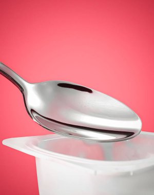 Kırmızı arka planda yoğurt kabı ve gümüş kaşık yoğurt kremalı beyaz plastik kap sağlıklı beslenme ve beslenme dengesi için taze süt ürünü.