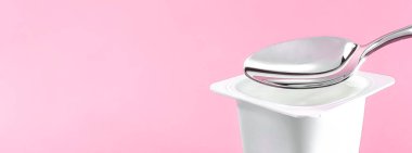 Yoğurt kabı ve pembe arka planda gümüş kaşık yoğurt kremalı beyaz plastik kap sağlıklı beslenme ve beslenme dengesi için taze süt ürünü.