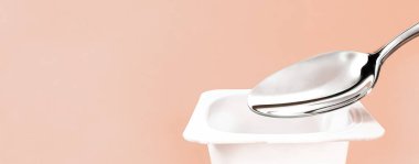 Yoğurt kabı ve bej arka planda gümüş kaşık, yoğurt kremalı beyaz plastik kap, sağlıklı beslenme ve beslenme dengesi için taze süt ürünü