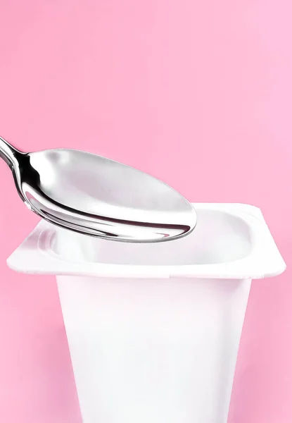 Tasse de yaourt et cuillère d'argent sur fond rose, récipient en plastique blanc avec crème de yaourt, produits laitiers frais pour une alimentation saine et l'équilibre nutritionnel — Photo