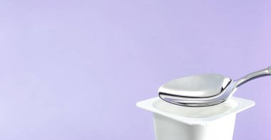 Yoğurt kabı ve mor arka planda gümüş kaşık yoğurt kremalı beyaz plastik kap sağlıklı beslenme ve beslenme dengesi için taze süt ürünü.