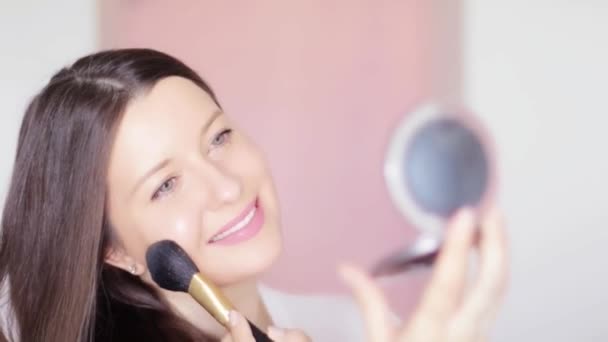 Mulher com escova de maquiagem e espelho compacto aplicando pó cosmético e sorrindo, rosto retrato de belo modelo em fundo rosa, ideia de maquiagem natural, cosméticos e produtos para a pele comercial — Vídeo de Stock