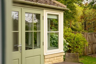 Garden room exterior showing door and window with reflections of green trees and plants in summer. Cotswolds countryside, England
