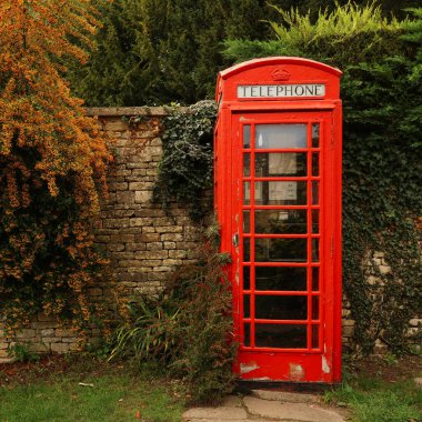 İngiliz kırsalında sonbahar yeşillikleriyle çevrili eski taş duvarın yanında duran kırmızı telefon kulübesi, tarihi iletişim sembolü. Cotswolds, İngiltere
