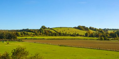 Countryside displaying undulating green hills, fertile pastures, and brown cultivated fields under a vibrant blue sky, highlighting farming and nature