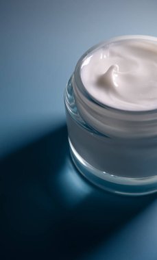 Şeffaf bir kavanozdaki kremsi beyaz nemlendirici, derin mavi bir yüzeye ince bir ışık tutar, su ve lüks güzelliği sembolize eder.