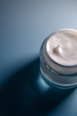 Beyaz, kalın, kremsi cilt ürünü şeffaf cam bir kavanozda, mavi bir arka planda, dokuyu gösteriyor.