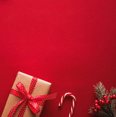 Kırmızı kurdeleli bir el işi kağıdına sarılı Noel hediyesi, bir şeker kamışı ve dokulu kırmızı arka planda böğürtlenli ladin dalı.