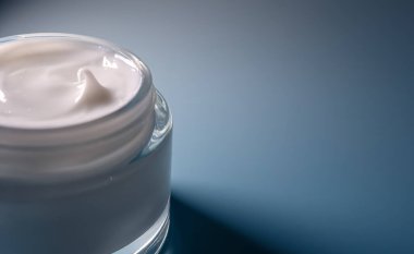 Beyaz cilt nemlendirici krem cam kavanozda, güzellik için kozmetik ürünleri nemlendiriyor ve mavi arka planda yaşlanmayı önlüyor.