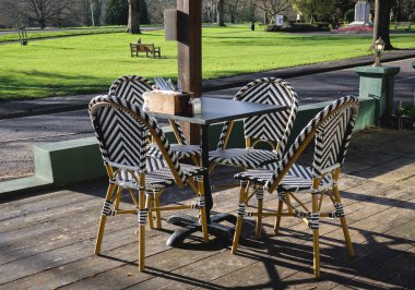 Açık kafeterya masası ve ahşap terasta sandalyeler, arka planda savaş anıtı ve sessiz parkı olan müşteriler için hazır.