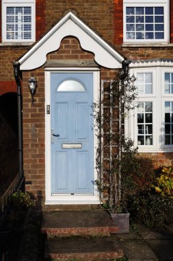 Açık mavi ön kapı ve beyaz gölgelik geleneksel tuğla ev cephesinde, hoş bir yerleşim girişi yaratıyor.