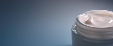 Açık cam bir kavanozda beyaz kozmetik krem, ince mavi arka planda pürüzsüz bir dokuyu gösteriyor, hidrasyon ve kendine bakım üzerine odaklanıyor.