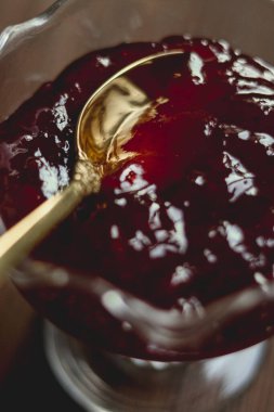 Kırmızı böğürtlen reçeli, altın bir kaşık, berrak bir cam kasede dinleniyor, tatlı ve zengin desenli bir meyve korunağı sergiliyor