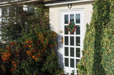 Evin ön kapısında çiçekli bir çelenk, etrafı bitkilerle çevrili ve uğurlu bir at nalı, evi ve kutlamayı simgeliyor.