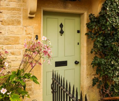 Kırsal taştan bir evin yeşil ön kapısında pembe çiçeklerle çevrili süslü siyah bir kapı tokmağı ve posta kutusu var.