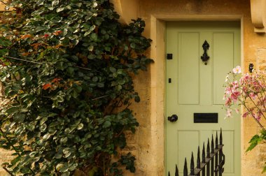 Kırsal taştan bir kulübenin geleneksel yeşil ön kapısı. Etrafı bol bitki yaprakları ve narin pembe çiçeklerle çevrili.