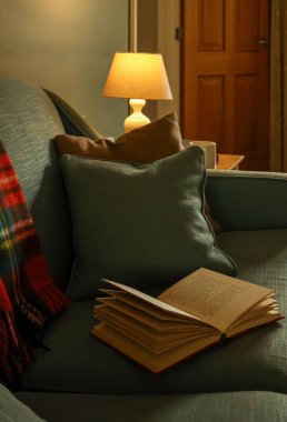 Battaniyeli kanepe ve sıcak ışıkta yastıklar, okunmuş ve gevşetilmiş sessiz bir akşamı davet eden açık bir kitap.