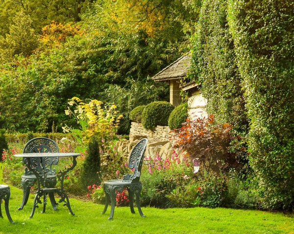 Outdoor seating area with a classic cast iron table and chairs, surrounded by lush plants, flowers, and an English cottage