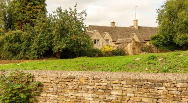 Geleneksel Cotswold taş bir duvarın ve bir elma ağacının arkasındaki kır evi İngiliz sükunetini gösteriyor.