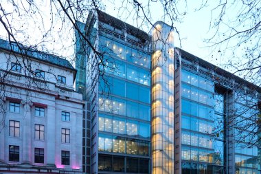 Londra bina cephesinde tarihi taş mimarinin yanında modern cam bir kule sergileniyor, alacakaranlıkta ağaç dallarından aydınlatılıyor.