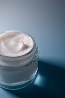 Yumuşak mavi arka planda açık cam bir kavanozda beyaz krem yüz ve vücut rutini için besleyici nemlendirici.