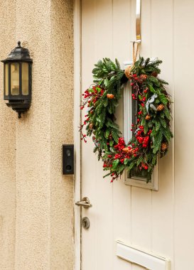 Krem rengi ön kapıda asılı Noel çelengi mevsimlik neşeyi yeşil yapraklar ve parlak kırmızı meyvelerle karşılıyor.