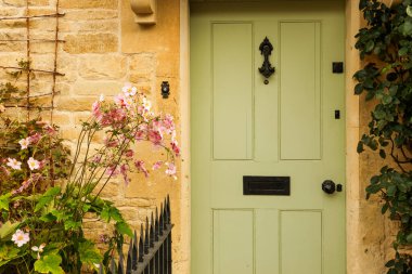 Yeşil bir ön kapısı, siyah tokmağı ve posta kutusu olan taşralı bir kır evi. Yeşillik ve canlı pembe çiçeklerle çerçevelenmiş. Büyüleyici, kusursuz bir ülke girişi için.