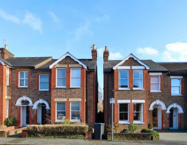 Açık mavi gökyüzünün altında beyaz süslemeli ve koyu pencereli geleneksel tuğlalı evler, İngiliz banliyölerinde yaşayan