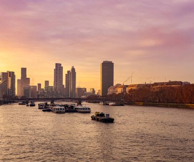 Günbatımında Londra şehir manzarası, Thames Nehri, modern gökdelenler, bir köprü ve sakin sularda çeşitli tekneler.