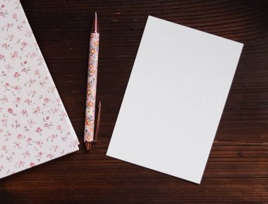 Boş kart ve çiçek desenli kalem. Koyu ahşap bir masanın üzerinde, pembe gül desenli bir defterin yanında. Yazmaya hazır.