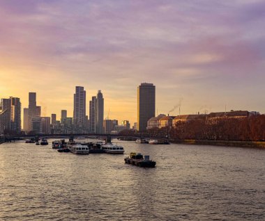 Londra ufuk çizgisi Thames Nehri 'ni gün batımında teknelerle donatıyor. Canlı turuncu ve mor renkler akşam gökyüzünü dolduruyor.