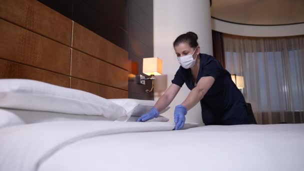 Trabajadora enmascarada arreglando cama en habitación de hotel — Vídeo de stock