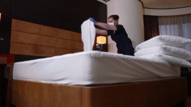 Eldivenli maskeli hizmetçi otel odasında yatak yapıyor.