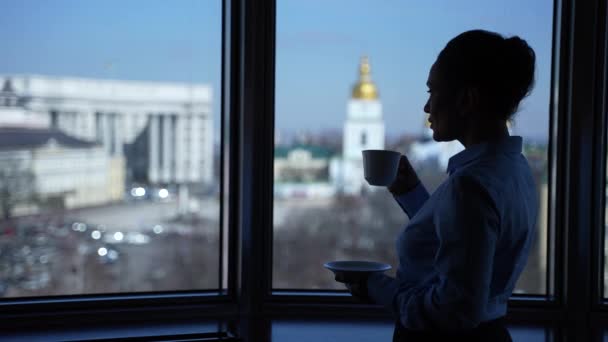 Egy nő kávét iszik, kinézve az irodai ablakon. — Stock videók