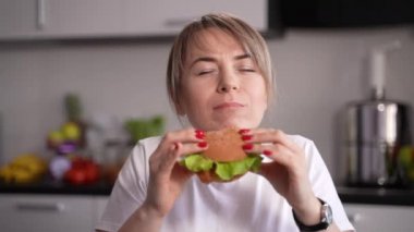 Mutfakta hamburger yiyen mutlu bir kadın.