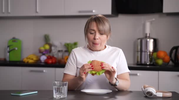 空腹の女性食べるハンバーガーキッチンに座って — ストック動画
