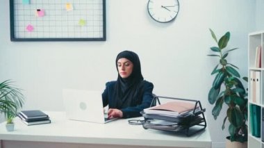 Görevdeki evrakların yanında dizüstü bilgisayar kullanan Müslüman iş kadını. 