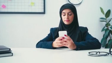 Masanın yanında akıllı telefon kullanan Müslüman bir iş kadını. 