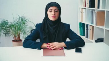 Müslüman iş kadını, kağıt klasör ve akıllı telefonun yanındaki kameraya bakıyor 